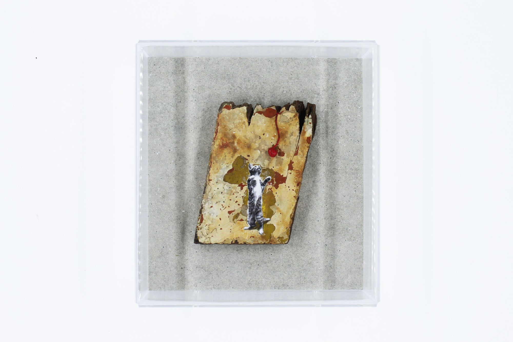 Small rectangular piece of stone with a painted cat and red ball of string on a gray background