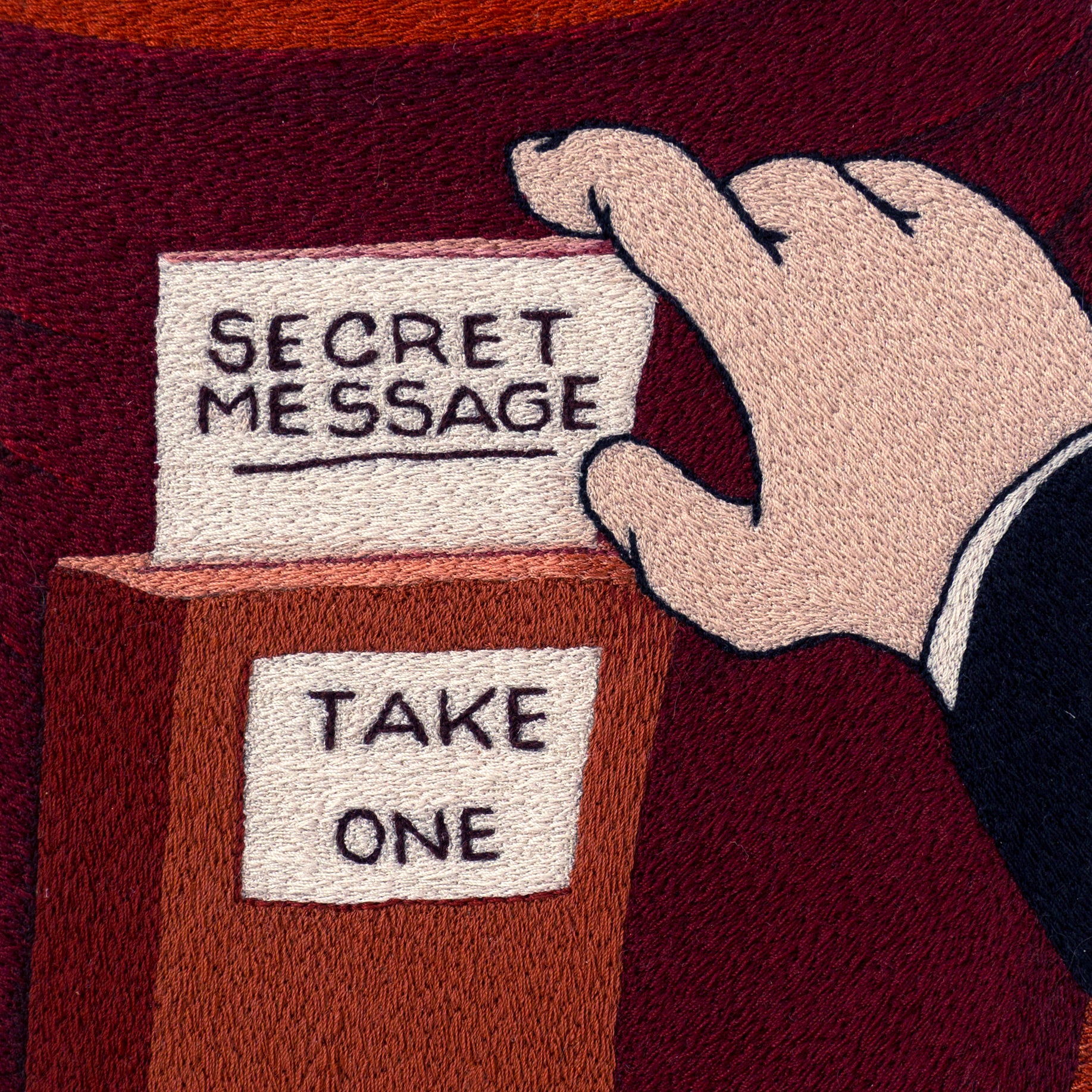 detail of an an embroidery of a hand taking a secret message from a box that says take one